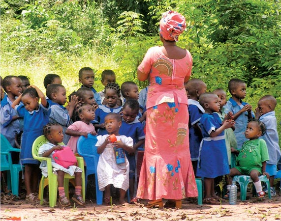 A Nigerian teacher trained by Rettig teaches an outdoor class. Students sit on plastic chairs that keep fire ants away from them while they learn. The chairs were purchased with funds that Rettig raised.