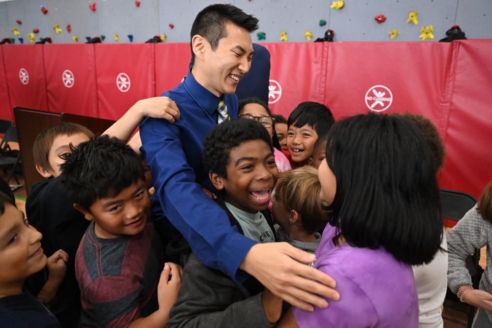 A man in a blue shirt smiles while being enthusiastically hugged by a group of children in a room with red mats and a climbing wall. This heartwarming scene features Shane Baker, a dedicated teacher from Kentucky who always brings joy to his students.